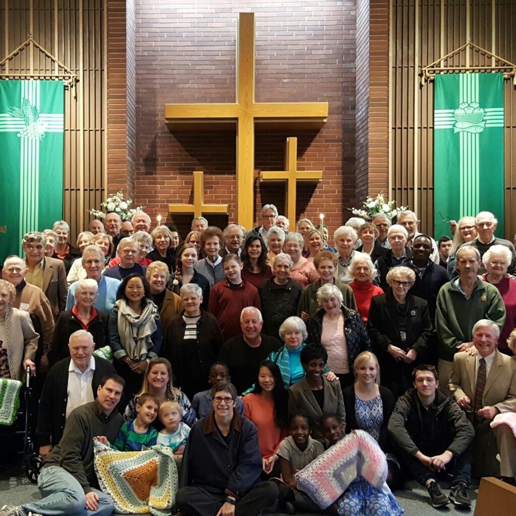 A large group of people, including diverse age groups, pose together inside a church. They stand in front of a wooden cross with green banners on either side. Some are seated; children hold colorful blankets. The setting suggests a community gathering.