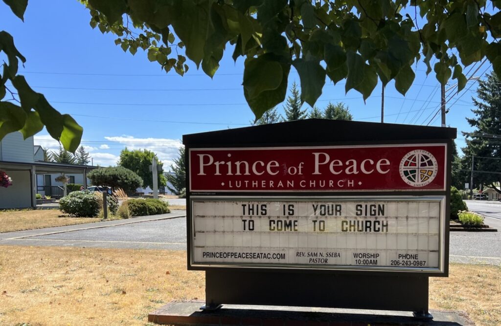 A sign for Prince of Peace Lutheran Church stands outdoors under tree branches. The sign reads, "THIS IS YOUR SIGN TO COME TO CHURCH," along with contact information and worship details. The sky is clear with a few clouds.