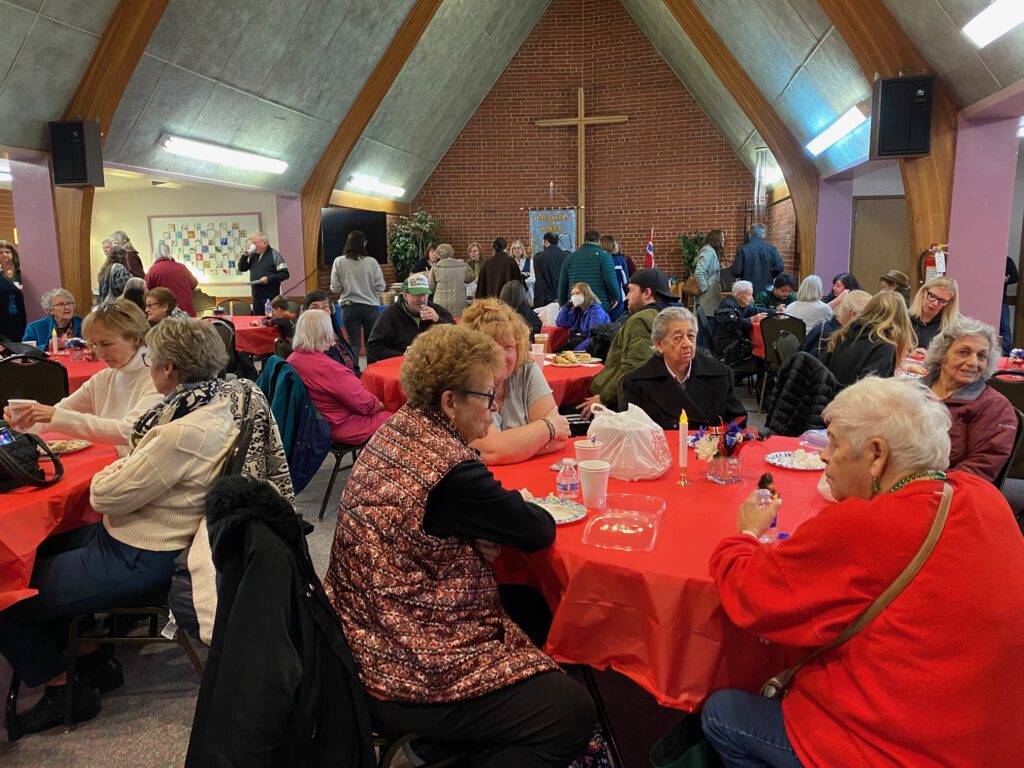 A large group of people sitting around tables with red tablecloths in a community hall with a cross on a brick wall. The room is full of lively conversation and activity, with attendees of various ages enjoying food and drinks.