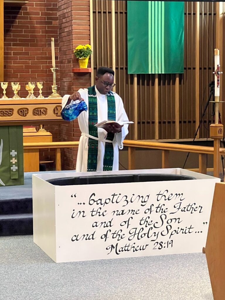 A pastor in a white robe and green stole pours water into a baptismal font. Behind them is an altar with a wooden cross and green banners. A quote from Matthew 28:19 is displayed on the font.