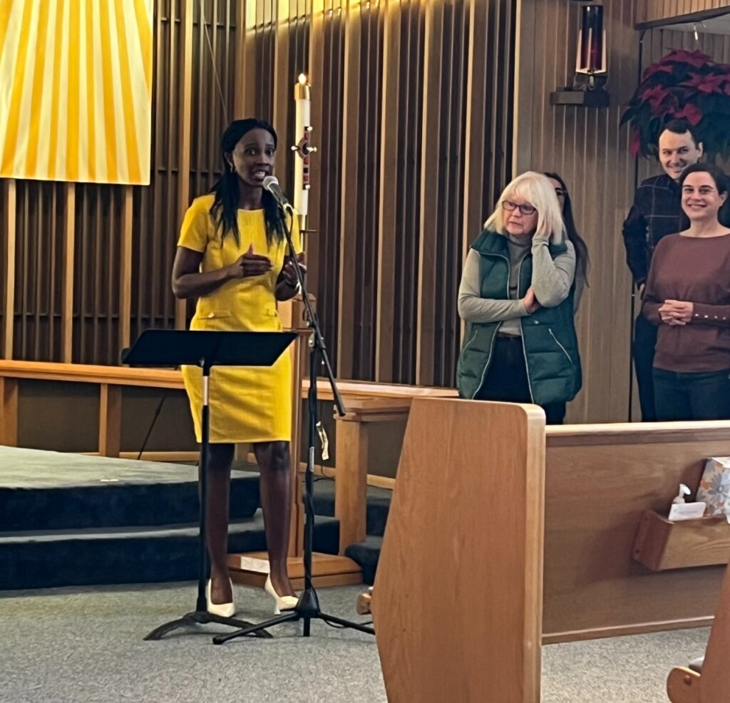 A woman in a yellow dress speaks at a microphone in a church or auditorium. Three people stand nearby, listening. The background features wooden paneling and a candle.