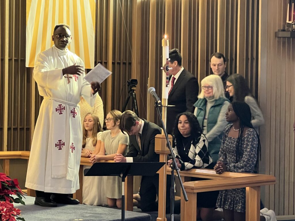 A pastor in a white robe stands at an altar holding papers, speaking to a congregation. Behind him, a group of people sit and stand, some holding programs. A lit candle is on the altar, and poinsettias decorate the area.