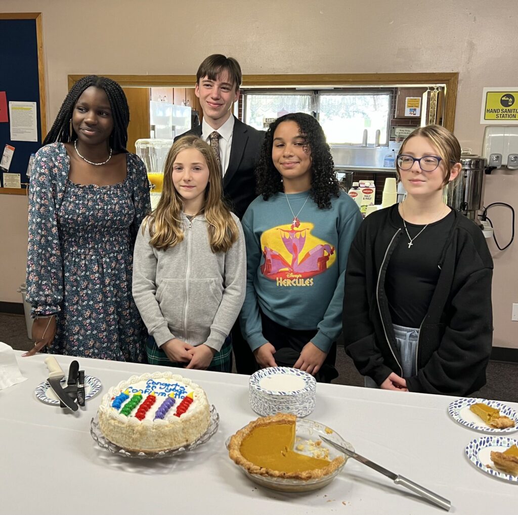 Five people stand behind a table with a cake and pumpkin pie. The cake has colorful decorations. They are in a room with beige walls and a window in the background.