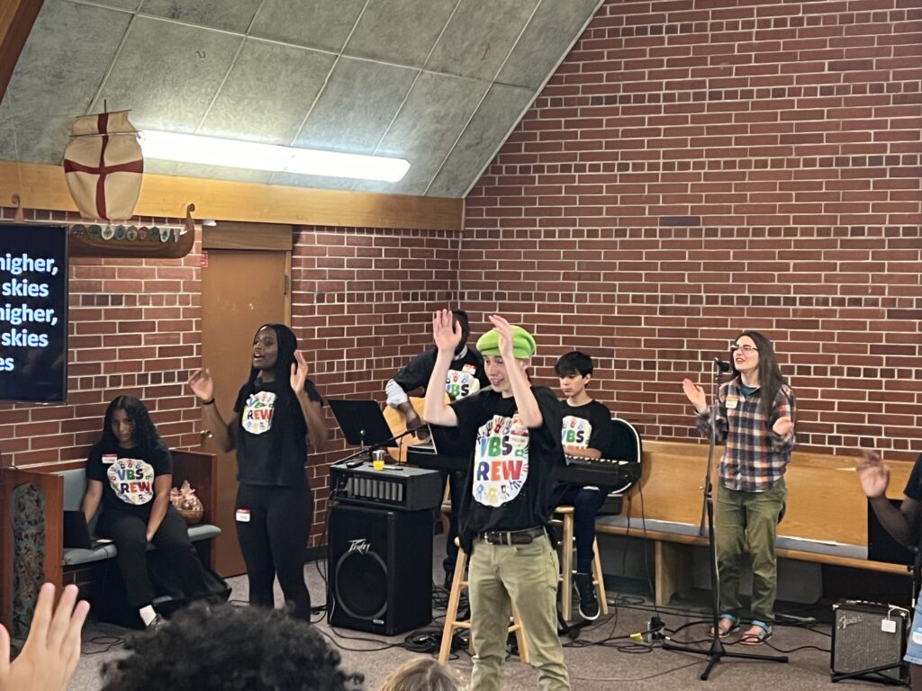 A group of people stand and sing in a brick-walled room during a performance. Some are clapping, some play instruments, and there are lyrics projected on the wall behind them. They wear matching black T-shirts with colorful text.