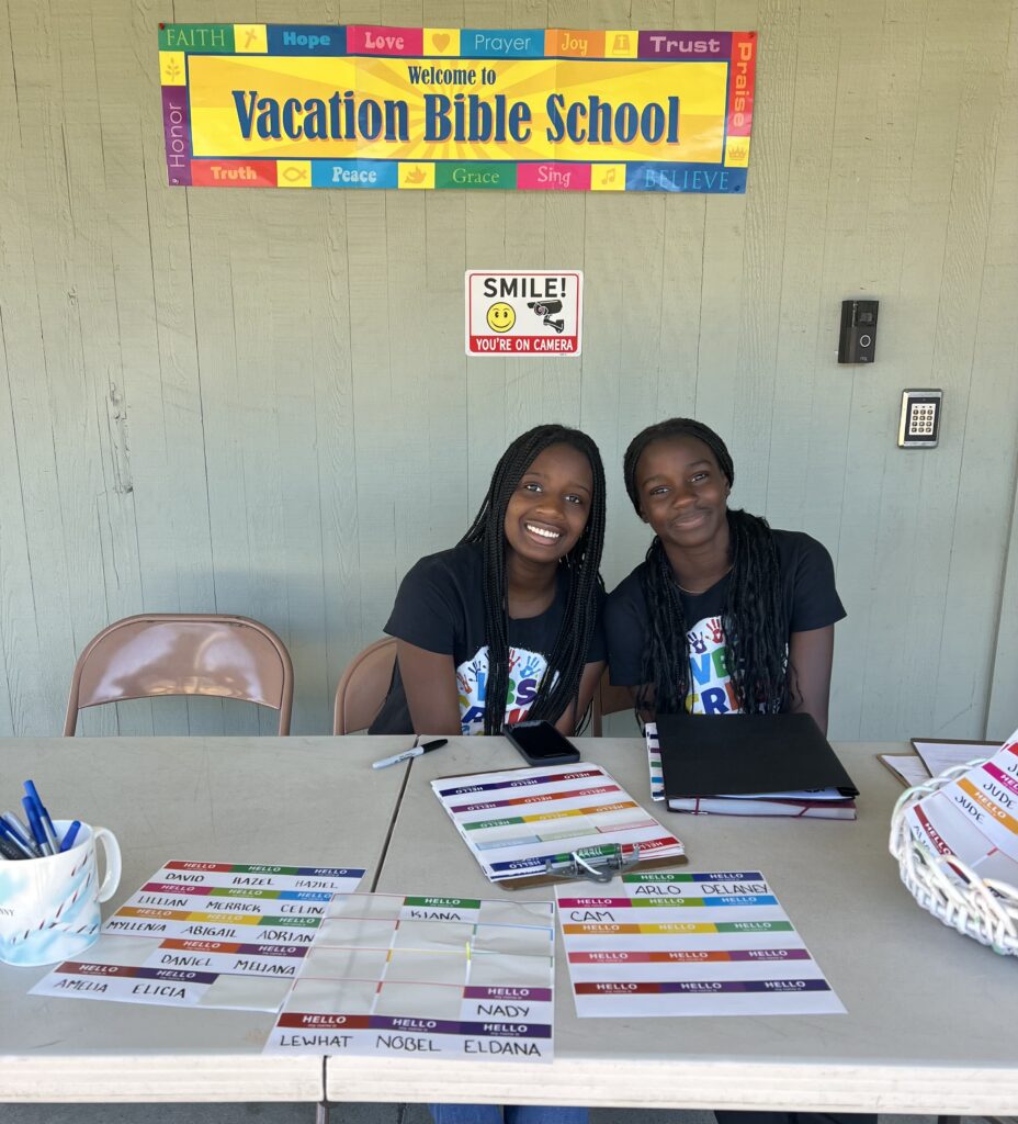 Two smiling people sit at a registration table for Vacation Bible School. The table has labeled name tags and pens. A colorful banner above reads "Welcome to Vacation Bible School,” with words like "Hope," "Love," and "Trust.