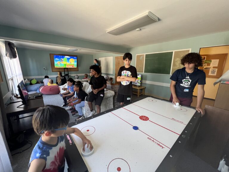 A group of young people are inside a recreation room. Two of them are playing air hockey while others are sitting on chairs and couches, focused on a video game displayed on a large screen. The room has a relaxed, casual atmosphere.