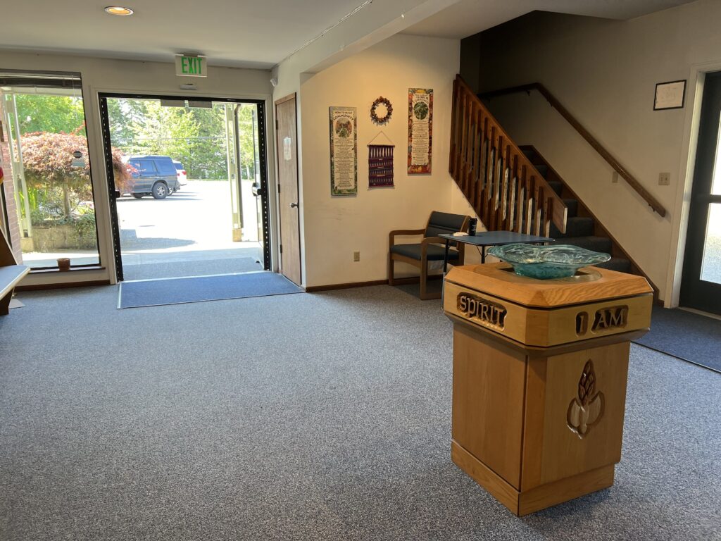 A serene foyer with an open entrance leading to a parking lot. A wooden baptismal font with "Spirit" and "I AM" is in the foreground, stairs in the background. Chairs and wall art decorate the space, offering a welcoming atmosphere.