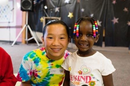 Two children smiling at the camera. The child on the left wears a tie-dye shirt and has short hair. The child on the right has colorful beads in their braids and wears a white t-shirt. They are indoors with a blurred background decorated with stars.