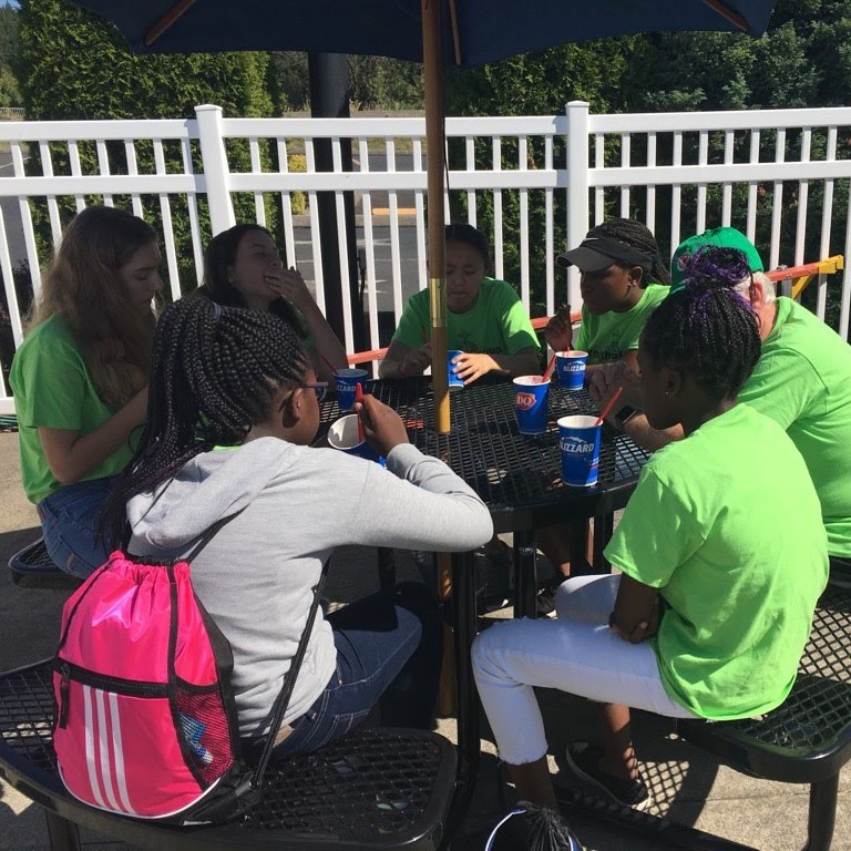 A group of people wearing green shirts sit around a picnic table under an umbrella, enjoying drinks. They appear to be engaged in conversation, with white fencing and greenery in the background.