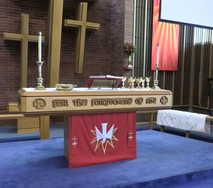 A church altar with a wooden table inscribed "For the Forgiveness of Sin." The altar is adorned with candles, chalices, and a red cloth featuring a white cross. A wooden cross and red banner are displayed in the background.